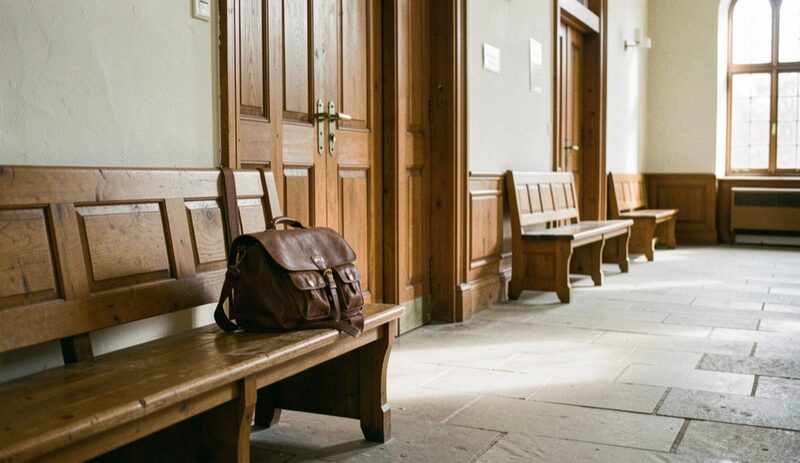Pasillo vacío de un edificio judicial con bancos de madera junto a la pared y altas puertas cerradas, una cartera de cuero marrón sobre uno de los bancos y luz de mañana entrando por los ventanales sobre el suelo de piedra.